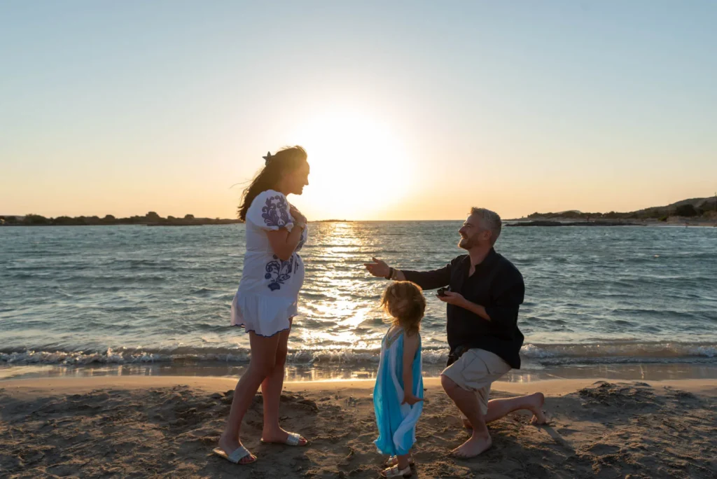 Zane kneeling to propose to Elsie at Elafonissi Beach in Crete during sunset with their daughter standing between them